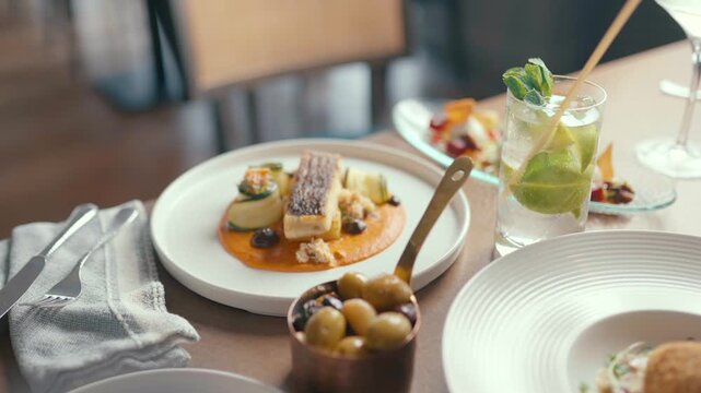 Elegant plated fish dish with sauce and olives on wooden table. Fresh green drink with mint beside main course. Soft focus background with chairs. Mood feels calm and luxurious