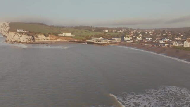 Aerial view of coastal cliffs and calm sea. Gentle waves wash along shore. Small town nestled at water's edge. Buildings line beachfront with green hills behind