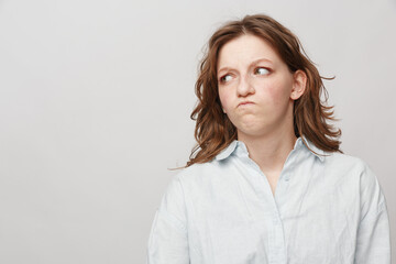 Fototapeta premium Young woman in white shirt looks sideways with skeptical expression on light background. Concept of doubt, suspicion, and uncertain emotions in casual portrait.