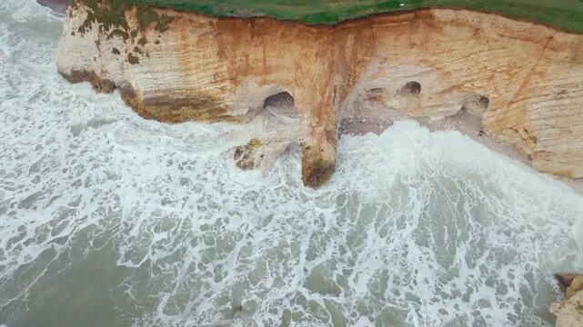 Aerial view of dramatic coastal cliffs with crashing ocean waves. Waves crash against layered rock formations, creating white foam. Green grass covers cliff tops, contrasting with sandy tones