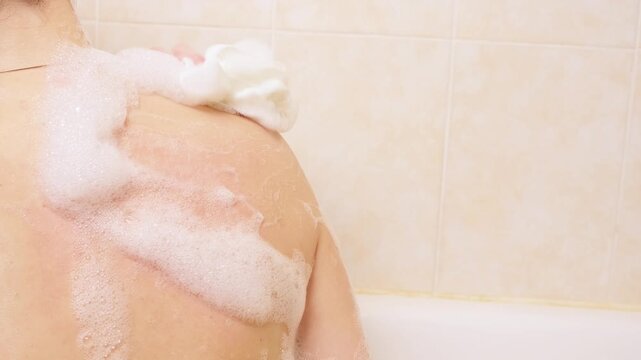 Woman washing shoulder with foamy sponge in bathtub. Beauty skin care and body cleansing routine in relaxing home spa environment.