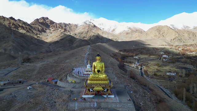A cinematic drone orbit captures the towering golden seated Buddha statue as it overlooks the rooftops of Stok, with the dramatic snow-covered peaks of the Ladakh Range stretching across the horizon.