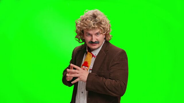 Comedic man with curly blond wig and mustache in brown blazer and patterned tie stands gesturing with hands, looking at camera on vivid neon green chroma key background plain