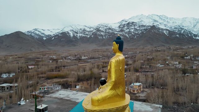 A cinematic drone orbit captures the towering golden seated Buddha statue as it overlooks the rooftops of Stok, with the dramatic snow-covered peaks of the Ladakh Range stretching across the horizon.