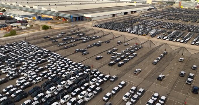 Aerial view of a large asphalt parking lot belonging to an industrial site, densely packed with rows of newly manufactured dark and light cars waiting for transport or distribution.