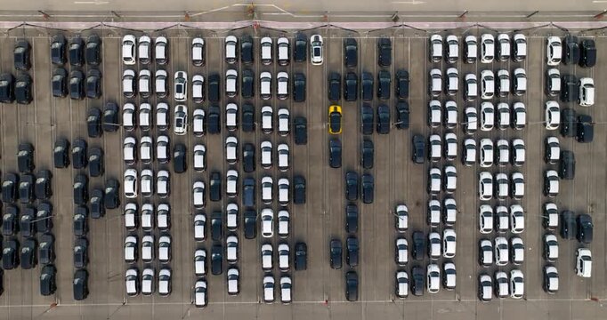 Zenithal aerial view captures a massive parking lot filled with hundreds of new cars organized in neat rows. The grid-like pattern features mostly black and white vehicles, creating a striking visual.