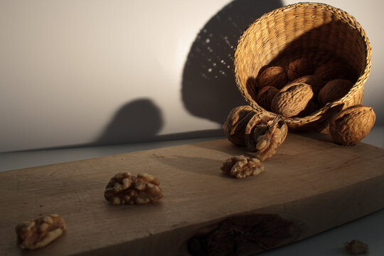 Walnuts spilling / falling out of a wicker basket onto a natural wooden board / still-life / still life / shadows / off-white background