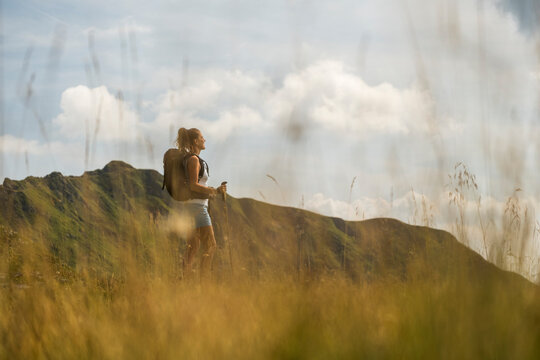 A woman with a backpack hikes in a grassy field, looking towards a mountain. Monte Crostis,Carnia,Friuli Venezia Giulia,Italy