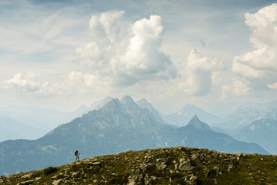 A hiker with a backpack and trekking poles walks on a rocky, grassy ridge, with mountains and a cloudy sky in the background. Monte Crostis,Carnia,Friuli Venezia Giulia,Italy