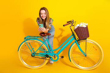 Young stylish woman with turquoise bicycle and basket of flowers smiles at her phone against a...