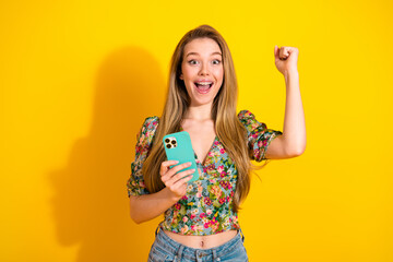 Happy young woman with phone celebrates in bright yellow studio wearing floral top and jeans