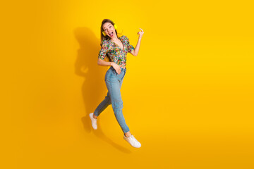 Young stylish woman in floral top jumping with headphones against bright yellow background for...