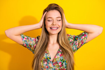 Young woman in floral dress smiles with hands on head against bright yellow background