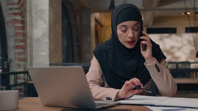 Muslim woman in hijab businesswoman discuss with phone business stats laptop review in office multitasking concentrated Islamic girl female manager consulting talking checking data in computer papers