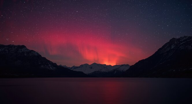 Brilliant red aurora shining above dark mountainous terrain and quiet water under night sky