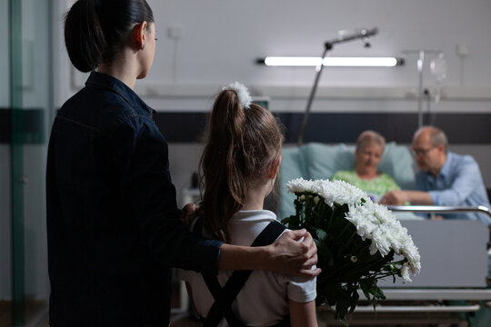 Young woman taking young daughter to visit recovering grandmother in hospital. Female relatives arriving, presenting flowers bouquet to elderly bedridden female patient at medical clinic.