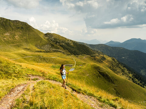 A woman hikes uphill on a grassy trail with trekking poles, mountains in the background. Monte Crostis,Carnia,Friuli Venezia Giulia,Italy