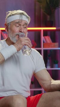 Leisure concept where likable smiling sportive bearded 70-aged man in sportswear replenishing water balance in his body after home workout and watching amusing messages drom friends on mobile