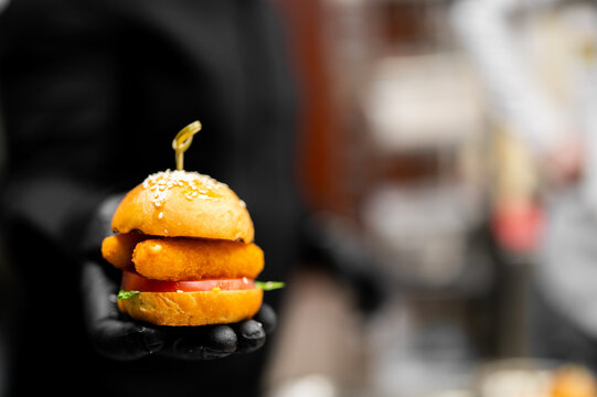 Close-up of a hand in a black glove holding a mini chicken burger slider with sesame bun, tomato, and lettuce on a blurred background.