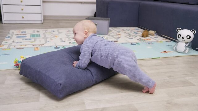 Cute baby in a knitted romper suit learning to climb over a large floor pillow, developing motor skills and coordination while playing on a colorful mat in the living room at home