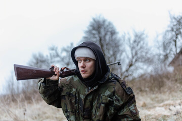 Fototapeta premium A male hunter with a gun in a camouflage jacket in a field in cold weather