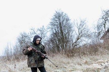 Fototapeta premium A male hunter with a gun in a camouflage jacket in a field in cold weather