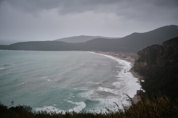 Fototapeta premium Wide scenic view of sweeping coastal bay where sea waves roll toward long sandy shoreline beneath dark overcast clouds. Mountainous hills and rugged cliffs frame coast, creating moody natural seascape