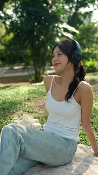 Young beautiful woman wearing headphones in casual style listening to music on smartphone while relaxing in a green city park, female enjoying peaceful nature and sunshine in modern urban lifestyle.
