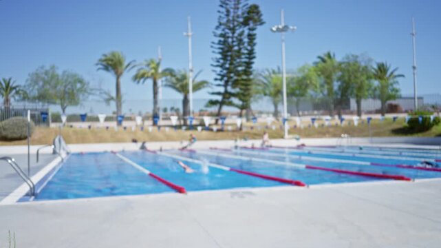 Blurred swimming pool scene with soft bokeh, defocused lane ropes, shimmering blue water and indistinct palm trees outdoor; background backplate copyspace calm.
