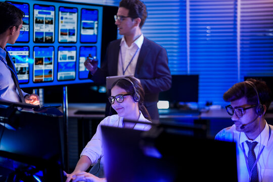 Female IT helpdesk technician wearing headset providing technical customer support during night shift. Professional dispatch operator managing client communications in dark neon office environment.