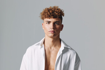 Obraz premium Young man with curly hair wearing open white shirt posing against gray background, looking confidently at camera with neutral expression and relaxed posture.