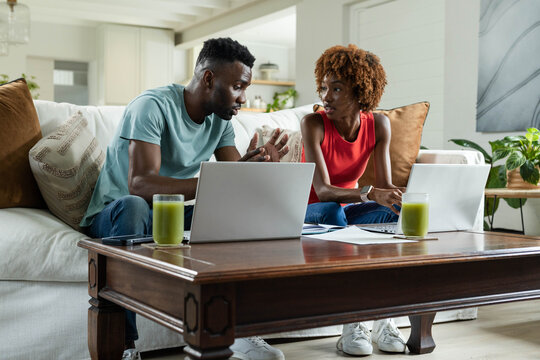 Couple in teal tee and red top sitting on sofa working laptops and paperwork with smoothies