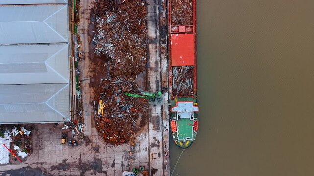 Scrap metal barge at dock. A barge is docked by a warehouse, loading scrap metal from the pile on the shore. This scene shows industrial activity.