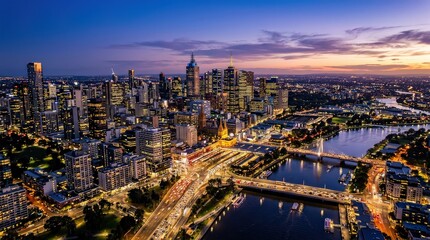 Obraz premium A vibrant cityscape at dusk showcases illuminated skyscrapers, busy roads, and a winding river under a colorful sky.