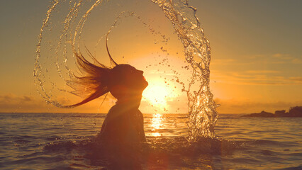 SILHOUETTE: Playful woman splashing ocean water by whipping her head backwards.
