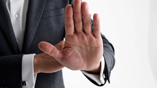 A professionally dressed individual, wearing a sharp suit and a crisp white shirt with elegant cufflinks, extends an open palm directly towards the viewer in a clear and decisive "stop" gesture. This 