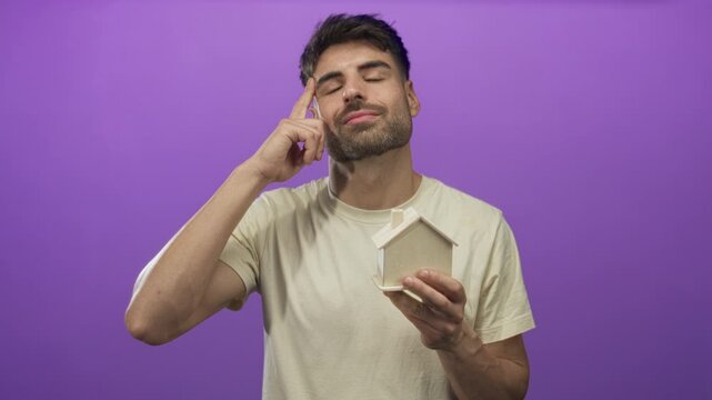 Young hispanic man holds wooden house model and points finger in purple studio; investment growth optimism.