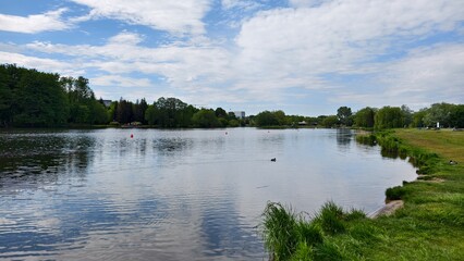 Willows grow on the grassy shore of the lake. There is a park on the shore, and behind it there are buildings and cranes. There are ripples and ducks on the water and buoys installed. Sunny spring © Balser