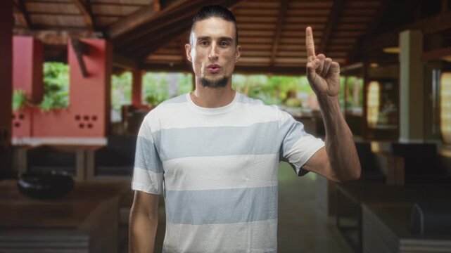 Man raising fist and waving open hand in a building, wearing striped tshirt and goatee while pointing finger; confidence greeting.