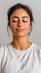 Fototapeta premium Young woman with freckles closes her eyes, meditating peacefully indoors. She wears a simple white t-shirt and delicate layered silver necklaces. Her face reflects a serene and calm state