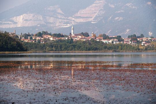 View of Annone Lake with calm water reflections, a small Italian village and church tower along the shore, and mountains rising in the background under a soft hazy sky.