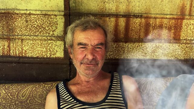 Portrait of homeless adult man sitting on worn couch placed and smoking outdoor. The setting underscores neglect and impermanence, presenting a raw and unfiltered glimpse of difficult living situation