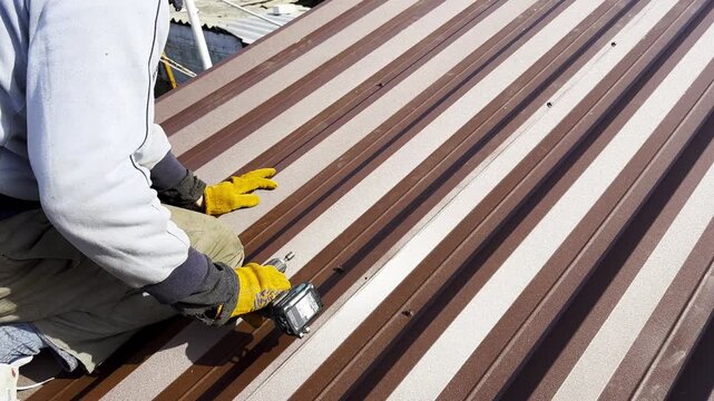 Male hands in gloves use cordless screwdriver to fasten roofing sheets in house roof. Young worker secures screws into surface performing careful installation work during residential construction