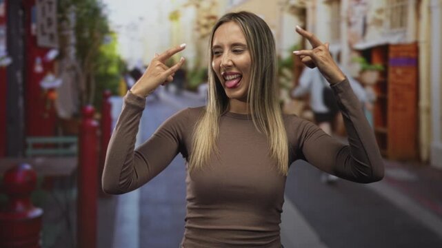 Woman showing rock horns with tongue out on street with both hands raised, smiling broadly; youthful playful.