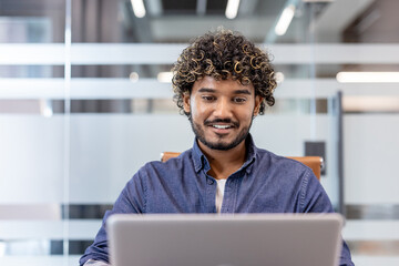 Close-up photo of a young Indian man smiling while working on a laptop in the office