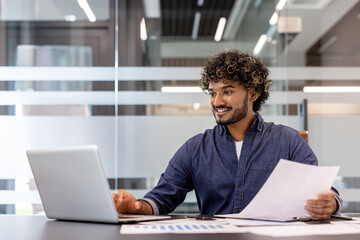 Smiling young Indian man working in the office with documents, holding papers in his hands and typing on a laptop