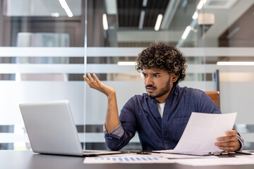 Problems with documents and data. A stressed young Indian man sits at a desk in an office and looks at a laptop screen