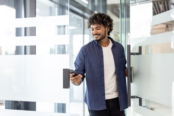 An Indian smiling man holds a phone in his hands, opens the door and enters the office premises