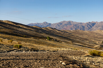 A wide view of the dry Bolivian highlands, showing sunlit brown hills leading to craggy mountain ranges under a clear sky.