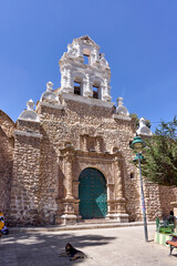 A view looking up at the San Benito church in Potosi. The church features a stone facade, a white bell tower, and an ornate green door. It is a sunny day.